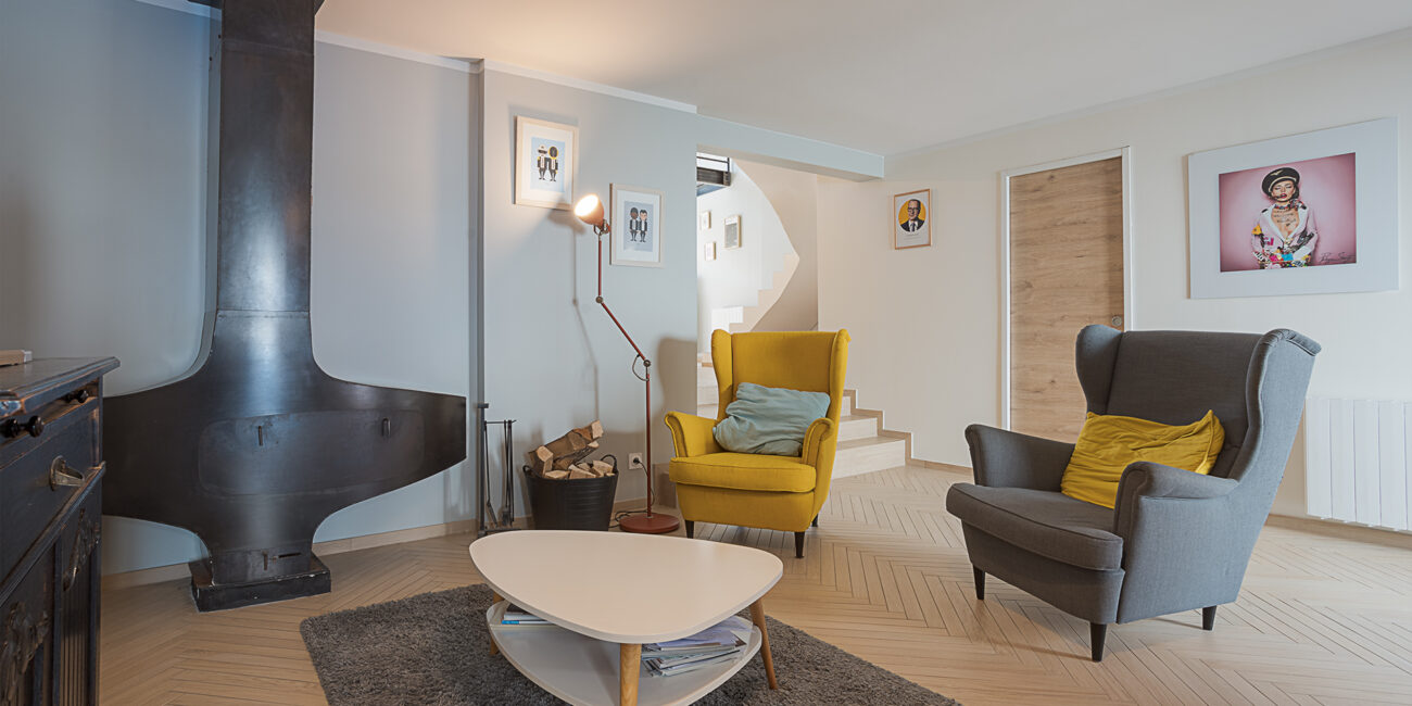 Photographie immobilière en Haute-Savoie, à Publier. photographie d'un salon avec une décoration moderne et colorée. on y voit deux canapé , un jaune un gris, une cheminé originale. Et des affiches colorées sont accrochées aux murs.